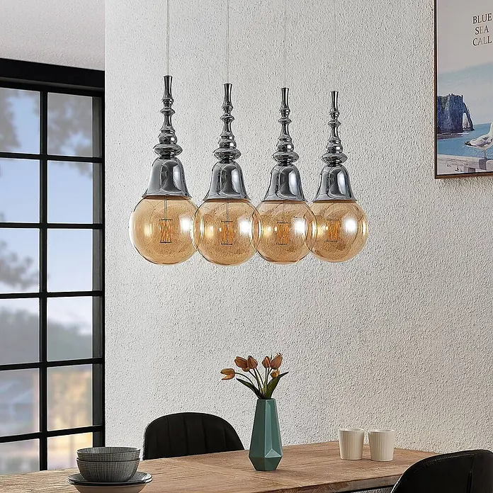 Lucande Pendelleuchte Gesja - L x B x H: 73,5 x 13,5 x 32,5 cm, chrom, E27 - Vier Pendelleuchten aus Chrommetall und bernsteinfarbenem Glas hängen über einem Esstisch und beleuchten den Raum.