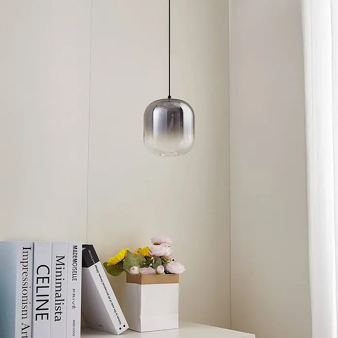 Lucande Pendelleuchte Avelinra - L x B x H: 14 x 14 x 17 cm, alu / grau / zink, rauchgrau, Schwarz - Eine moderne Pendelleuchte aus rauchfarbenem Verlaufsglas hängt von der Decke über einem Regal mit Büchern und Blumen.
