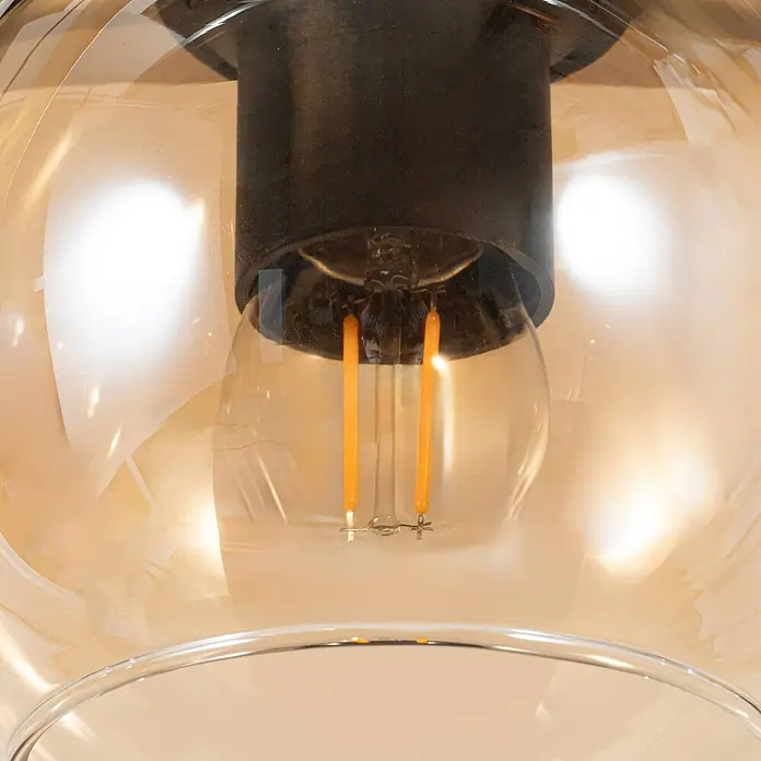 Lindby Pendelleuchte Enrique - creme / amber, amber, Schwarz, eiche hell, E27 - Nahaufnahme beleuchteter Lampe: schwarzer Sockel, bernsteinfarbener Glasschirm, LED-Glühlampe.