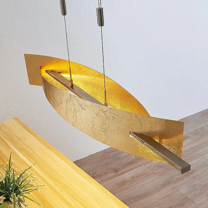 Lucande Pendelleuchte Marija - L x B x H: 101 x 18,5 x 1,8 cm, messing / gold, gold, silber - Goldfarbene Pendelleuchte mit geschwungenem Design und strukturierter Oberfläche, hängt über einem Holztisch.
