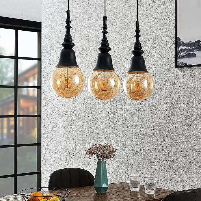 Lucande Pendelleuchte Gesja - L x B x H: 63,5 x 13,5 x 32,5 cm, Schwarz, E27 - Drei schwarze und bernsteinfarbene kugelförmige Pendelleuchten über einem Holzesstisch.