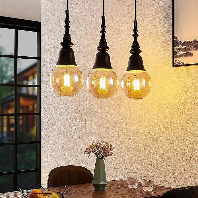 Lucande Pendelleuchte Gesja - L x B x H: 63,5 x 13,5 x 32,5 cm, Schwarz, E27 - Drei schwarz-bernsteinfarbene Pendelleuchten aus Glas und Metall über einem Esstisch.