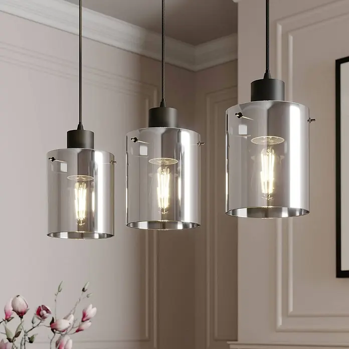 Lindby Pendelleuchte Kourtney - alu / grau / zink, Schwarz, rauchgrau, E27 - Drei Pendelleuchten mit schwarzem Sockel und zylindrischen Klarglasschirmen, die Edison-Glühbirnen in einem modernen Raum zeigen.
