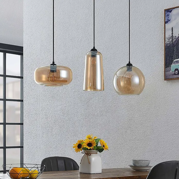 Lucande Pendelleuchte Wilja - creme / amber, Schwarz, amber, E27 - Drei Pendelleuchten mit bernsteinfarbenen Glasschirmen über einem Esstisch.