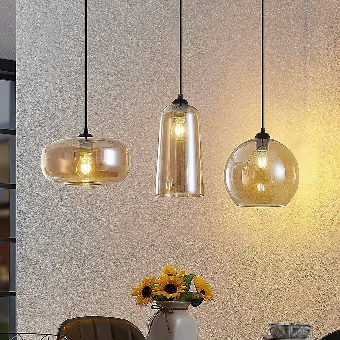 Lucande Pendelleuchte Wilja - creme / amber, Schwarz, amber, E27 - Drei Pendelleuchten aus bernsteinfarbenem Glas mit sichtbaren Glühbirnen, jede in einzigartiger Form, hängen über einem Tisch und beleuchten eine texturierte Wand.