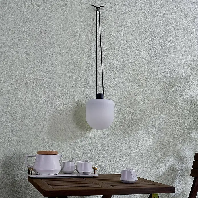 Lindby Außenwand- & Deckenleuchte Eleia - weiß / opal, IP44 - Weiße mattierte Glas-Pendelleuchte mit schwarzem Kabel über einem Holztisch mit Teeservice hängend.