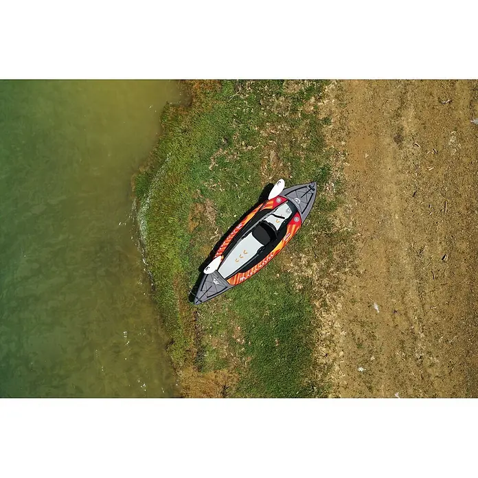 Aqua Marina Kajak Memba 10,10" - L x B: 330 x 90 cm, Geeignet für: 1 Person, Tragkraft: 110 kg - Ein orange-graues Schlauchkajak mit weißem Paddel liegt am grasbewachsenen Ufer neben grünlichem Wasser.
