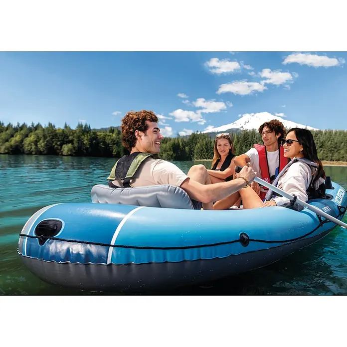 Intex Schlauchboot-Set Excursion 4 - Geeignet für: Max. 4 Personen, L x B x H: 315 x 165 x 43 cm - Vier Personen in einem blau-grauen Schlauchboot auf einem See, umgeben von grünen Bäumen und einem schneebedeckten Berg.