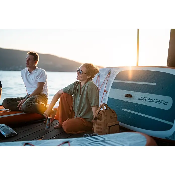 Aqua Marina SUP-Board-Set Pure Air Tropic 12" - L x B x H: 366 x 84 x 15 cm, Aufblasbar, Mehrfarbig, Tragkraft: 170 kg - Zwei Personen entspannen auf aufblasbaren Paddleboards am Wasser, mit einem braunen Rucksack neben ihnen bei Sonnenuntergang.