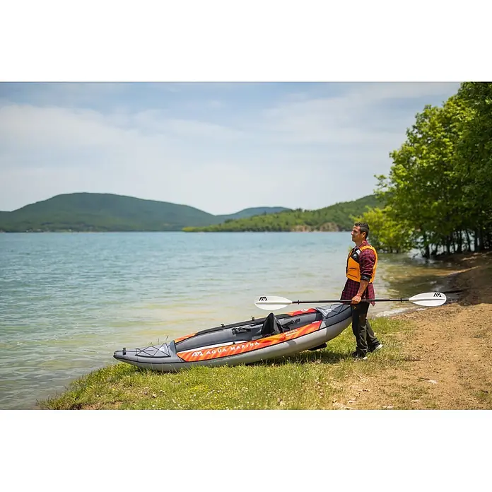 Aqua Marina Kajak Memba 10,10" - L x B: 330 x 90 cm, Geeignet für: 1 Person, Tragkraft: 110 kg - Ein Mann trägt ein grau-orangefarbenes Schlauchkajak und Paddel zu einem See.