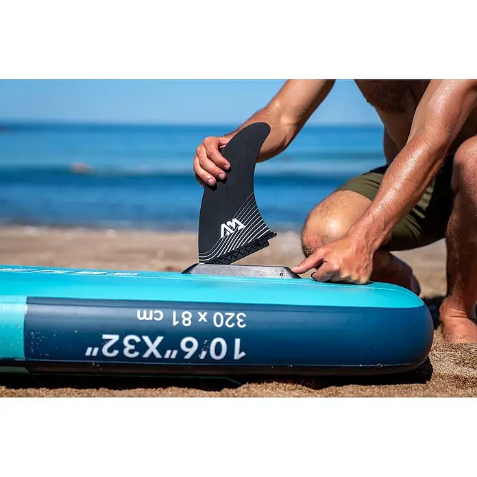 Aqua Marina SUP-Board-Set Beast 10,6" - L x B x H: 320 x 81 x 15 cm, Mehrfarbig, Tragkraft: 140 kg - Eine Person befestigt eine schwarze Finne mit weißen Markierungen an einem türkisfarbenen aufblasbaren Paddleboard am Strand.