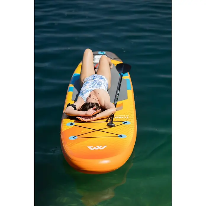 Aqua Marina SUP-Board-Set Turbo 330 - L x B x H: 320 x 81 x 15 cm, Mehrfarbig, Tragkraft: 140 kg - Ein orange-gelbes Stand-Up Paddleboard schwimmt auf blauem Wasser, eine Frau entspannt sich darauf, daneben liegt ein Paddel.