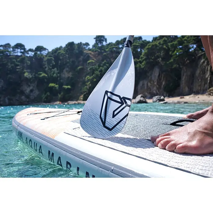 Aqua Marina SUP-Paddel Solid - 1 Stk., 180 cm - 220 cm - Nahaufnahme von Füßen auf einem weiß-orangen Stand-Up-Paddleboard in klarem Wasser, mit Paddelblatt sichtbar.