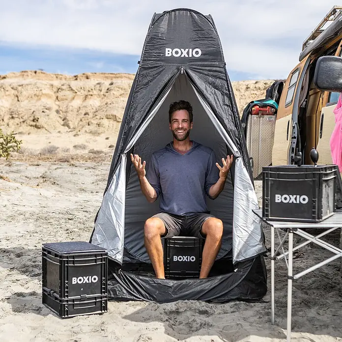 Boxio Zelt Dusche & Toilette - L x B x H: 63,5 x 63,5 x 6 cm, Schwarz - Schwarzes BOXIO Pop-up-Privatzelt mit Mann auf BOXIO Toilettenbox, im Sand neben einem Van verwendet.