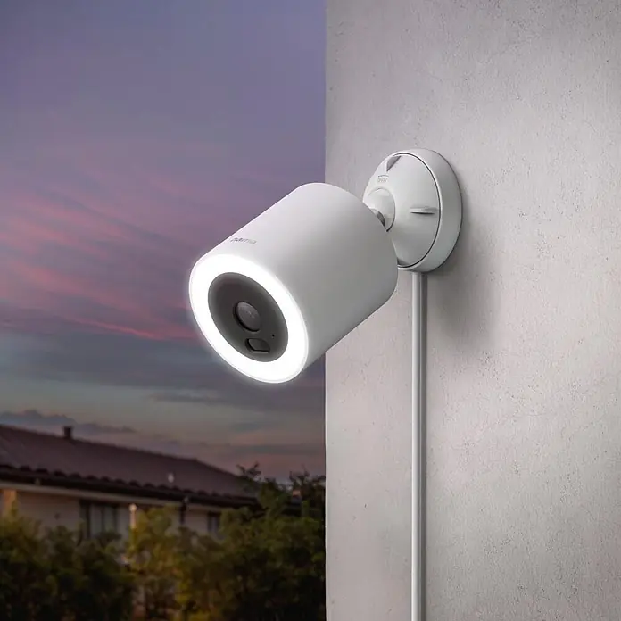 Hama Überwachungskamera 00176670 mit Licht u. Bewegungsmelder smart -  - Eine weiße Outdoor-Überwachungskamera mit beleuchtetem Ringlicht ist bei Dämmerung an einer grauen Wand montiert und überwacht ein Haus mit Bäumen.