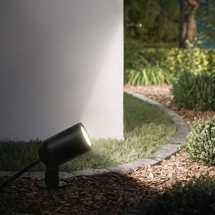 Hama LED-Gartenstrahler WLAN Gartenstecker - Schwarz - Schwarzer Außenstrahler beleuchtet eine weiße Wand in einem Garten mit Rindenmulch.