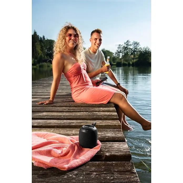 Hama Bluetooth-Lautsprecher 00188234 Bluetooth "Bomb 3.0" wasserdicht IPX7 LED 16 W -  - Schwarzer tragbarer Lautsprecher auf einem Holzsteg, neben einem pinkfarbenen Handtuch, mit Leuten am See.