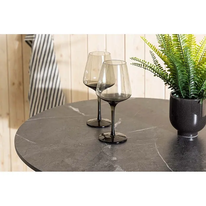 Ebuy24 Gartentisch rund Milano - Ø x H: 70 x 75 cm, Aluminium, Weiß, Stapelbar - Zwei rauchgraue Weingläser auf einem dunkelgrauen Tisch mit Marmorierung und einer Topfpflanze daneben.