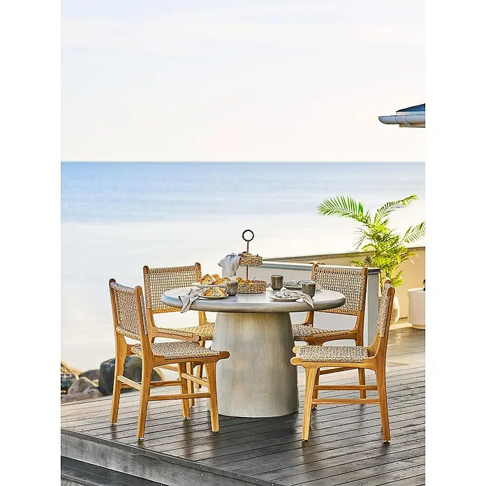 Ebuy24 Gartentisch rund Bente - Ø x H: 120 x 75 cm, 95% MgO, 5% Glasfaser, Grau, Stapelbar - Essgruppe für draußen mit rundem grauem Betontisch und vier Holzstühlen mit geflochtenen Sitzen und Lehnen, auf einer Terrasse am Meer.