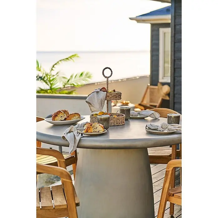 Ebuy24 Gartentisch rund Bente - Ø x H: 120 x 75 cm, 95% MgO, 5% Glasfaser, Grau, Stapelbar - Zweistöckiger Servierständer aus geflochtenen Naturfasern auf einem Gartentisch, beladen mit Obst und Gebäck.