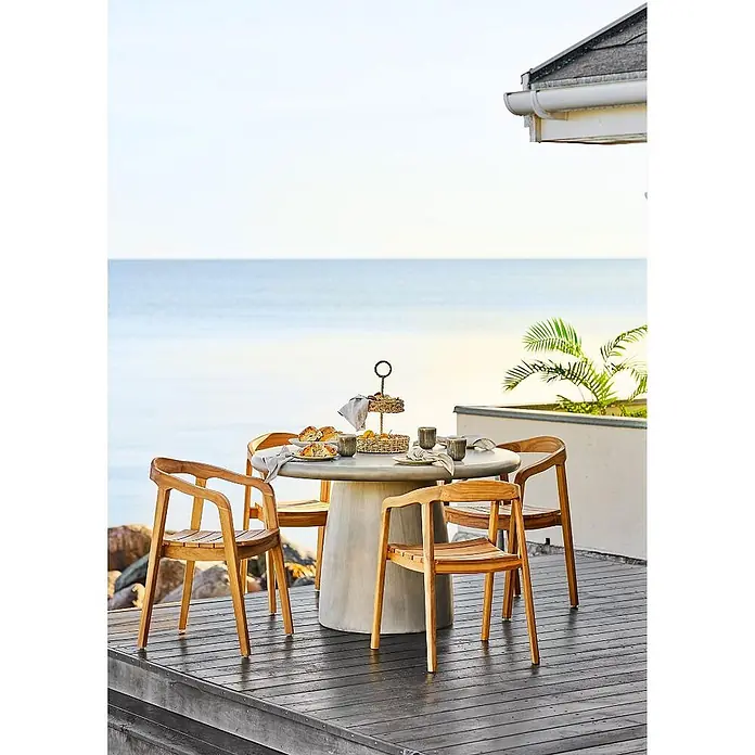 Ebuy24 Gartentisch rund Bente - Ø x H: 120 x 75 cm, 95% MgO, 5% Glasfaser, Grau, Stapelbar - Runder hellgrauer Betontisch mit vier Stühlen aus Naturholz auf einer Terrasse am Meer, gedeckt mit Etageren und Geschirr.