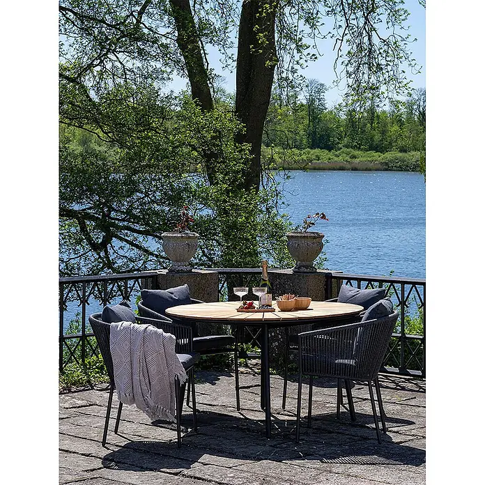Ebuy24 Gartentisch rund Cleveland - Ø x H: 130 x 74 cm, Teakholz Metall, Teak, Stapelbar - Runder Esstisch mit Holzplatte und dunklen Stühlen mit Kissen auf einer Terrasse am See, gedeckt für Getränke.