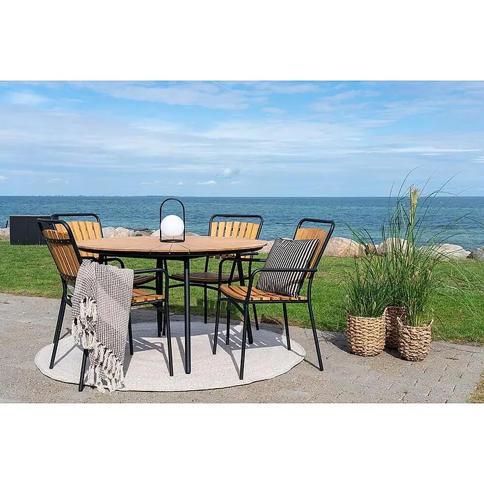 Ebuy24 Gartentisch rund Cleveland - Ø x H: 130 x 74 cm, Teakholz Metall, Teak, Stapelbar - Outdoor-Esstischgruppe mit rundem Holztisch und vier Stühlen mit schwarzen Metallrahmen und Holzlatten, auf einer Terrasse am Meer.