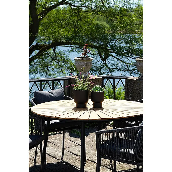 Ebuy24 Gartentisch rund Cleveland - Ø x H: 130 x 74 cm, Teakholz Metall, Teak, Stapelbar - Runder Esstisch für den Außenbereich mit Holzlattenplatte, schwarzen Beinen und schwarzen Geflechtstühlen mit Kissen, auf einer Terrasse.