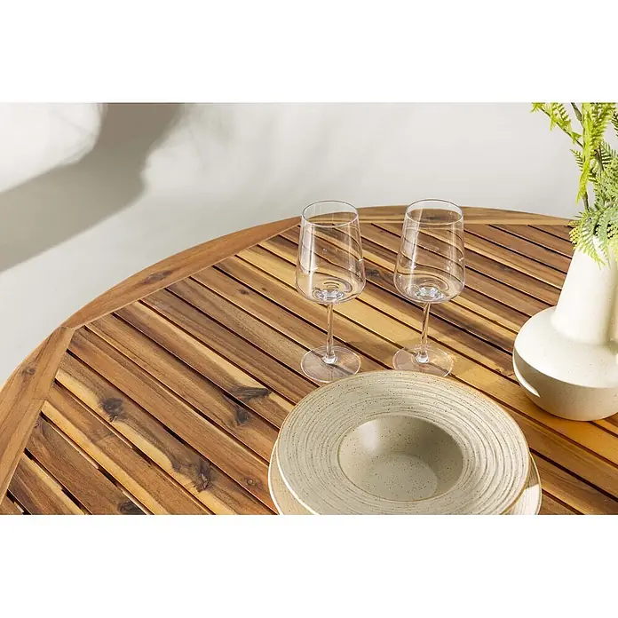 Ebuy24 Gartentisch rund Lumpur - Ø x H: 120 x 75 cm, Akazie, Natur, Stapelbar - Runder Holztisch mit Lattenstruktur, gedeckt mit Weingläsern, Tellern und einer Vase mit Grünpflanze.