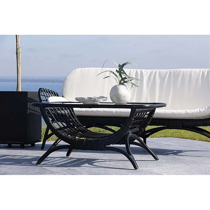 Ebuy24 Garten-Loungetisch Moana - Rattan, Schwarz, Stapelbar, L x B x H: 96 x 60 x 47 cm - Schwarzer Rattan-Couchtisch für draußen mit Glasplatte und passendem Sofa mit weißen Kissen auf einer Terrasse.