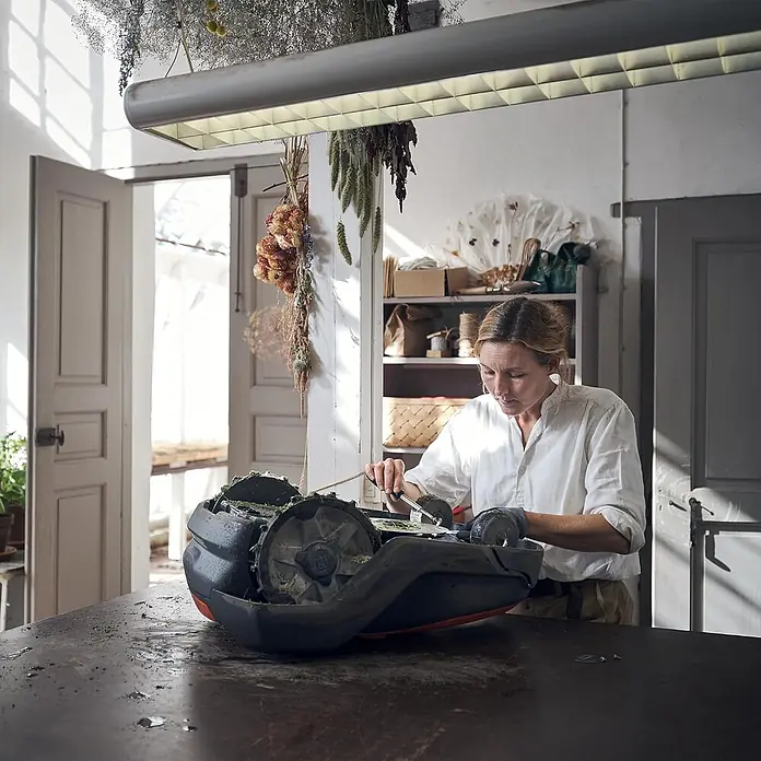 Husqvarna Ersatzmesser-Set für Mähroboter Classic Messer 45er-Pack - Passend für: alle Husqvarna Automower Modelle, 45 Stk. - Person reinigt einen grau-orangen Mähroboter und entfernt Gras von Rädern und Unterseite.