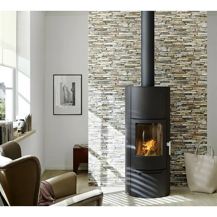 A.S. Création Papel pintado de vellón Wood-n-Stone - Beige/Marrón/Gris, Efecto piedra, 10,05 x 0,53 m - Elegante salón con una estufa de leña negra y moderna frente a una pared de piedra decorativa y un sillón cómodo.