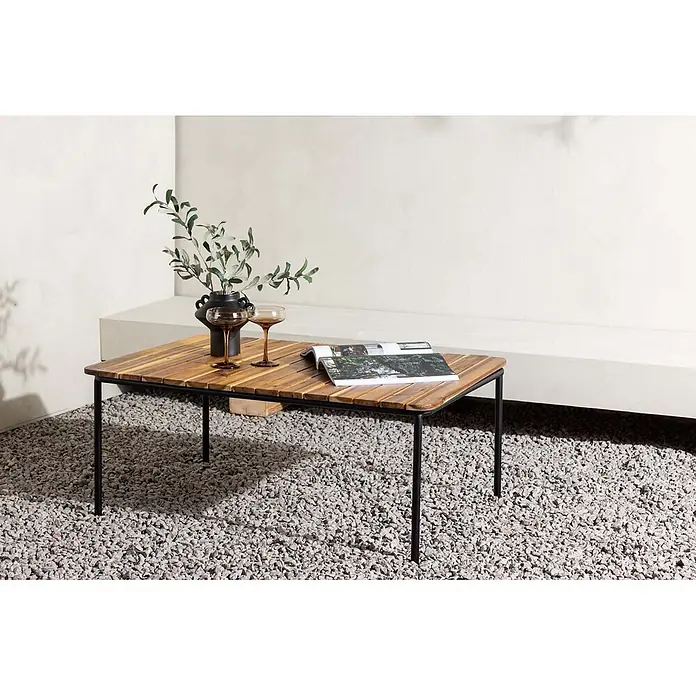Ebuy24 Garten-Loungetisch Penh - Akazie, Natur, Stapelbar, L x B x H: 100 x 65 x 40 cm - Outdoor-Couchtisch mit natürlicher Holzlamellenplatte und schwarzen Metallbeinen, auf einer Kiesterrasse gezeigt.