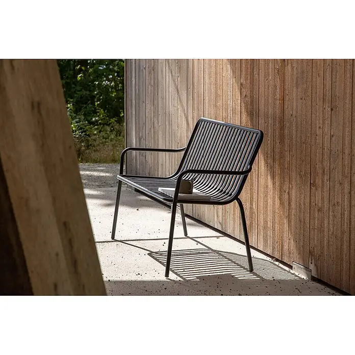 Ebuy24 Gartenbank Peking - B x T x H: 64 x 130 x 85 cm, Aluminum., Schwarz - Schwarze Metallbank mit Armlehnen, Buch und Tasse, steht draußen an einer Holzwand.