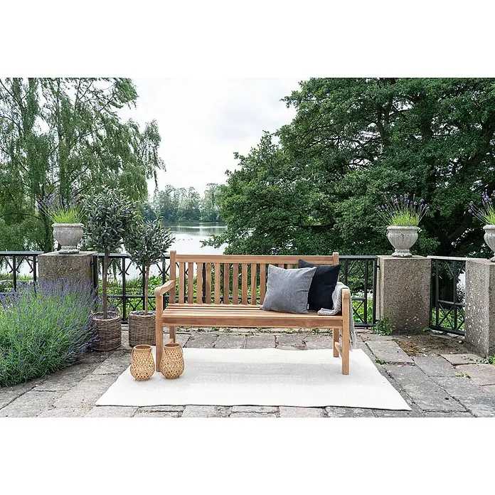 Ebuy24 Gartenbank Cabo - B x T x H: 150 x 150 x 92 cm, Teak., Holz - Hölzerne Gartenbank mit zwei Kissen und weißem Teppich auf einer Steinterrasse mit Seeblick.