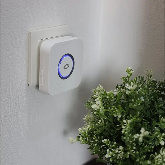 REV Funk-Gong-Set Piezo - Reichweite ca.: 100 m, 0 dB - 110 dB, IP20 - Weißer quadratischer Smart Plug mit blau beleuchtetem Ring und REV-Logo, in eine Wandsteckdose neben einer Grünpflanze gesteckt.