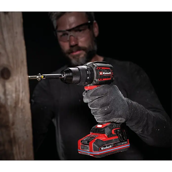Einhell Professional PXC 18V Akku-Schlagbohrschrauber TTP-CDD 18/501-C Li-i - 18 V, 1 Akku, 3 Ah, 50 Nm - Mann mit Schutzbrille und Handschuhen benutzt roten und schwarzen Einhell Akku-Bohrschrauber zum Schrauben in ein Holzbrett.