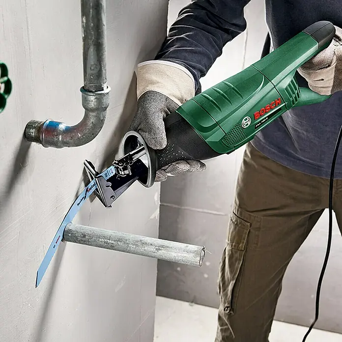Bosch Säbelsäge PSA 700 E - 710 W, Hublänge: 20 mm, 1 x Säbelsäge, 1 x 1 Sägeblatt für Holz S 2345 X - Eine grüne Bosch Säbelsäge in Aktion, die ein Metallrohr durchtrennt, getragen von einer Person mit Arbeitshandschuhen.