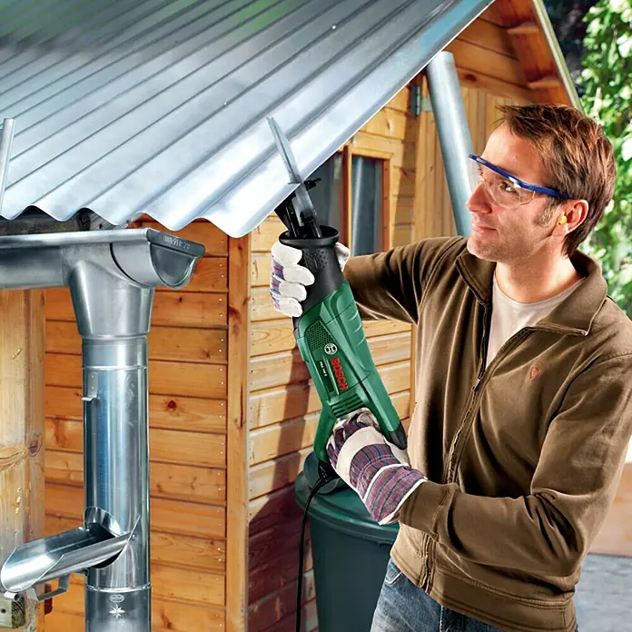 Bosch Säbelsäge PSA 700 E - 710 W, Hublänge: 20 mm, 1 x Säbelsäge, 1 x 1 Sägeblatt für Holz S 2345 X - Ein Mann benutzt eine Bosch Säbelsäge, um ein Dachrinnenrohr zu bearbeiten, wobei er Schutzkleidung trägt.