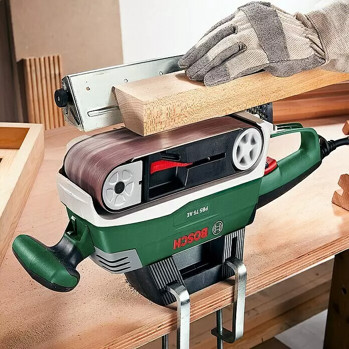 Bosch Lijadora de banda eléctrica de sobremesa PBS 75 AE - 750 W - Lijadora de banda Bosch verde y blanca fijada a un banco de trabajo, una persona lija una tabla de madera.