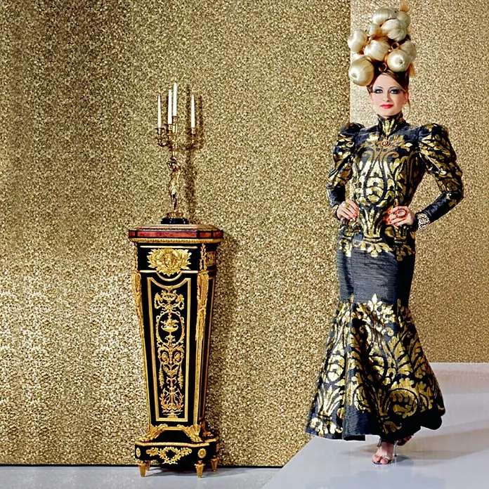 Glööckler Vliestapete - Federmuster - Gold, Ornament, 10,05 x 0,7 m - Frau in einem goldenen, verzierten Kleid neben einem goldenen, dekorierten Beistelltisch.