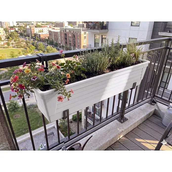 Plastard Pflanzkasten 100 cm selbstwässernd mit Hydroeinsatz & Metallhalterung - Außenmaß (L x B x H): 100 x 21 x 17 cm, Kunststoff, Weiß - Ein weißer rechteckiger Balkonkasten mit bunten Blumen und Kräutern, an einem schwarzen Geländer mit Stadtblick.