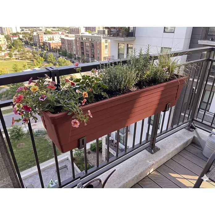 Plastard Pflanzkasten 3er Set 100 cm selbstwässernd Hydroeinsatz & Metallhalterung - Außenmaß (L x B x H): 60 x 21 x 17 cm, Kunststoff, Terrakotta - Brauner Kunststoff-Balkonkasten mit Blumen und Kräutern, an einem schwarzen Metallgeländer mit Stadtblick.