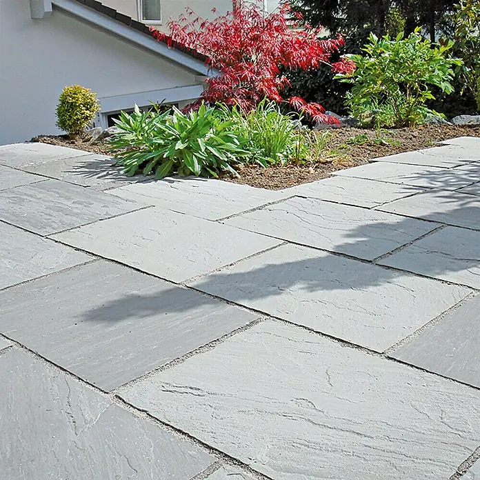 Terrassenplatte Delhi Grey - 30 x 30 x 2,5 cm, Grau, Sandstein - Graue Terrassenplatten aus Naturstein erzeugen einen modernen und eleganten Außenbereich mit Pflanzen und einem Haus im Hintergrund.