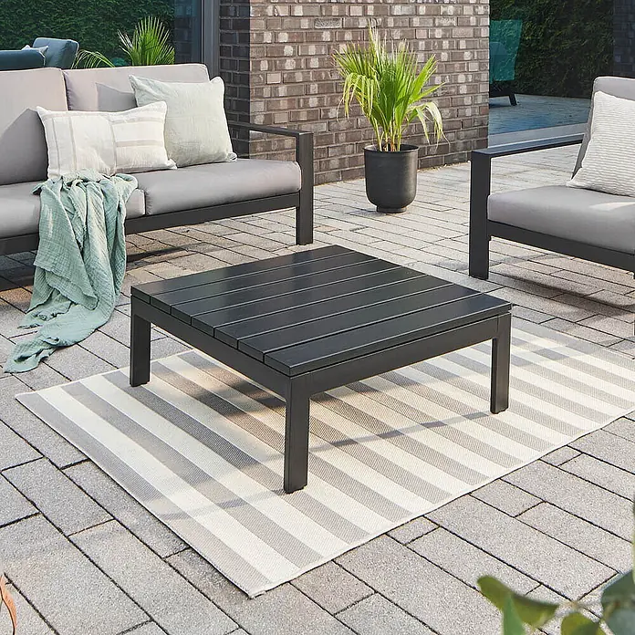 Home Deluxe Gartentisch Tischplatte RIO - L x B x H: 80 x 80 x 5 cm, Schwarz - Dunkelgrauer quadratischer Outdoor-Couchtisch mit Lattenrost auf gestreiftem Teppich als Teil eines Gartenmöbelsets.