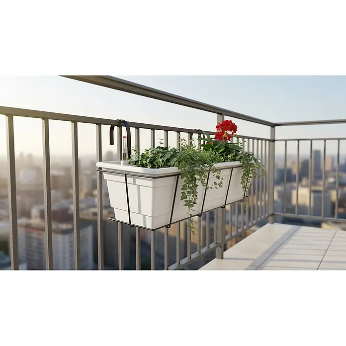 Plastard Pflanzkasten 100 cm selbstwässernd mit Hydroeinsatz & Metallkorb - Außenmaß (L x B x H): 100 x 21 x 17 cm, Kunststoff, Weiß - Weißer Balkonpflanzkasten mit schwarzer Metallhalterung, gefüllt mit Pflanzen und roten Blumen, an einem Geländer über einer Stadt.