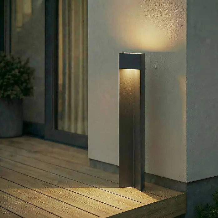 Lucande Außenlaterne Kaholo - 6 x 15 x 90 cm - Dunkelgraue moderne Pollerleuchte wirft warmes Licht auf Holzterrasse und helle Wand, schafft Outdoor-Ambiente.