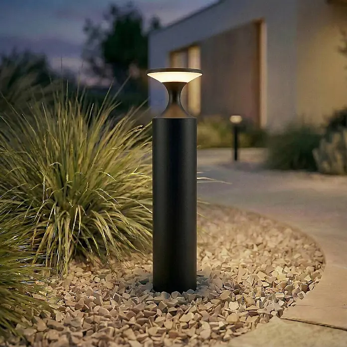Lucande Außenlaterne Navor -  - Schwarze Outdoor-Pollerleuchte mit warmem Licht, die einen Gartenweg mit Kies und Pflanzen beleuchtet.