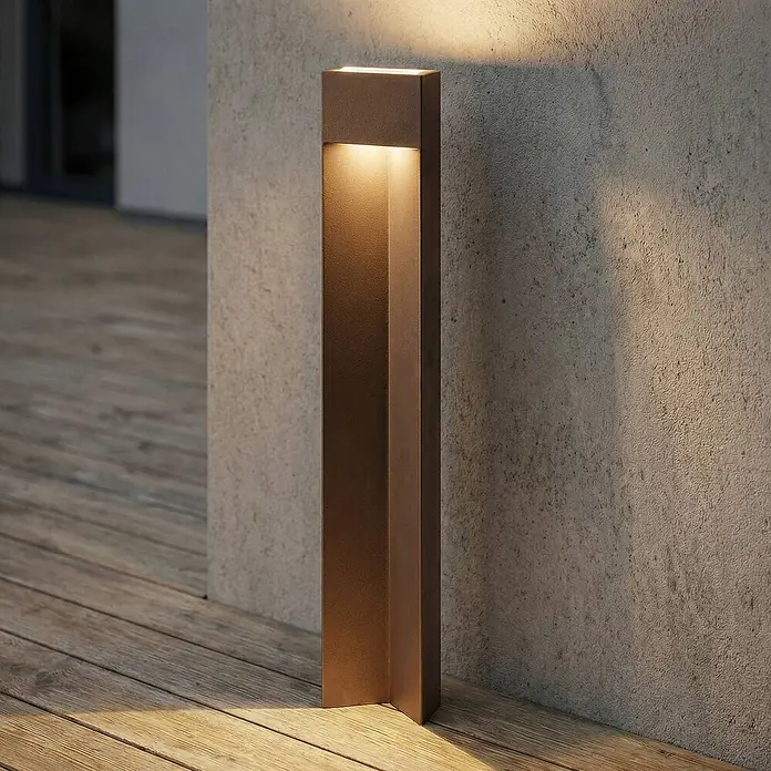 Lucande Außenlaterne Kaholo - 6 x 15 x 90 cm - Moderne bronzefarbene Pollerleuchte, die eine strukturierte Wand und Holzterrasse beleuchtet und warmes Licht nach oben und unten wirft.