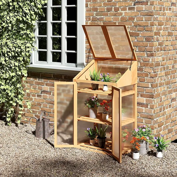Outsunny Gewächshaus - 70 x 50 cm, Material Seitenwand: Kunststoff, 4 mm, Braun - Holzkleinhaus mit transparenten Paneelen, offenem Dach und geöffneten Türen, gefüllt mit Topfpflanzen, an einer Ziegelwand.